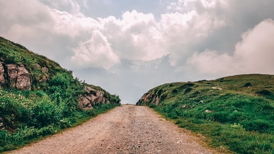 Under the white clouds and green grass field between brown dirt road
